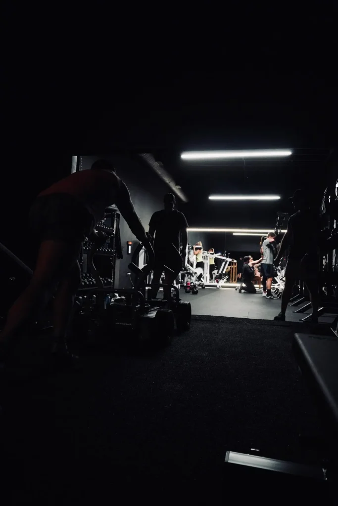 Dimly lit gym with multiple people engaging in strength training and pushing sleds under overhead lights.